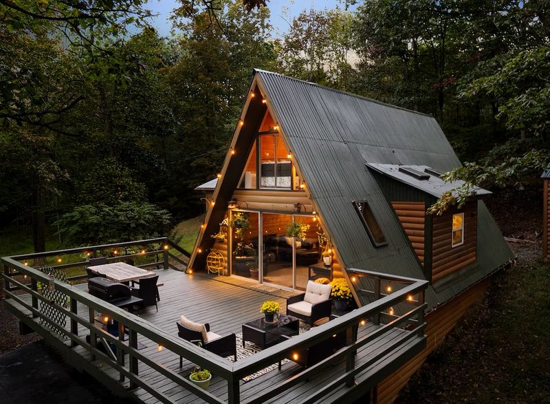 An A-frame mountain cabin lit up at dusk, with a furnished deck
